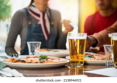 Close-up glass of beer with froth on table next to a plate with Margherita pizza. Unrecognizable group of people unfocused on background enjoy and eating. - Powered by Shutterstock