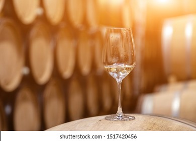 Closeup glass with white wine on background wooden wine oak barrels stacked in straight rows in order, old cellar of winery, vault. Concept professional degustation, winelover, sommelier travel