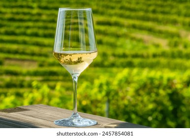 Close-up of a glass of white wine on a wooden table, with green vineyards in the background. Wine tasting, rating, and savoring - Powered by Shutterstock