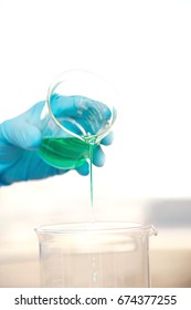 Closeup Of Gloved Hand Pouring Green Liquid Into Beaker Against Blurred Background