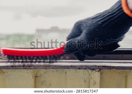 Closeup of gloved hand using red wire brush to clean metal balcony frame before painting, removing rust, flakes, and surface debris during outdoor home repair work with blurred city background