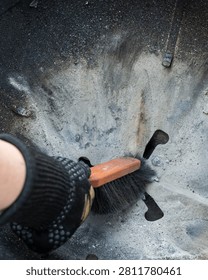 Close-up of a gloved hand using a small broom to sweep ash, soot and dirt from the inside of a kettle grill. The used barbecue surface shows gray ash, black grime and cleaning work after grilling. Suitable for barbecue, grill cleaning, maintenance, outdoor cooking, household chores and food preparation themes.