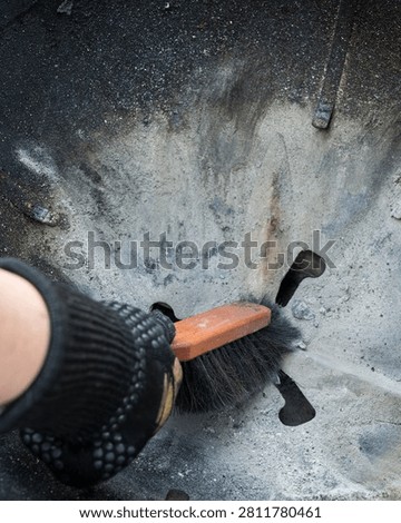 Close-up of a gloved hand using a small broom to sweep ash, soot and dirt from the inside of a kettle grill. The used barbecue surface shows gray ash, black grime and cleaning work after grilling. Suitable for barbecue, grill cleaning, maintenance, outdoor cooking, household chores and food preparation themes.