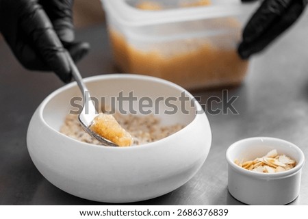 Close-up of gloved hands adding fruit compote to oatmeal in a white bowl. Healthy breakfast preparation with sliced almonds on stainless steel kitchen surface