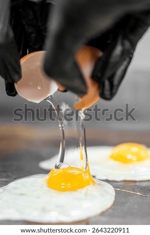 Close-up of gloved hands cracking a raw egg onto a hot surface, with yolk and egg white spreading during cooking process in a kitchen setting