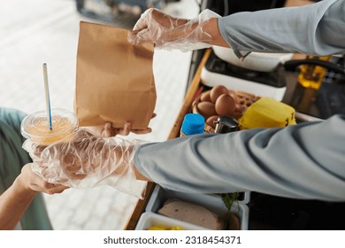 Close-up of gloved hands of truck vendor passing soda and packed fast food in paperbag over containers with ingredients for hotdogs - Powered by Shutterstock