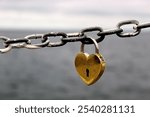 Close-up of a golden closed heart-shaped padlock hanging on a metal chain on the seashore. Padlock isolated on sea background.