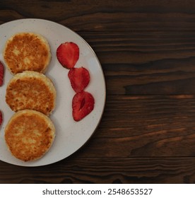 Close-up of golden-brown syrniki cottage cheese pancakes on a white plate with sliced strawberries, set against a rustic wooden surface. A cozy, traditional breakfast or dessert scene. - Powered by Shutterstock