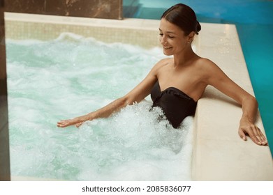 Close-up of a gorgeous elegant dark-haired woman in black swimsuit getting hydromassage at wellness spa resort. Middle aged beautiful brunette smiles enjoying hydro therapy sitting in a thermal pool