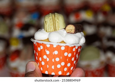 A close-up of a gourmet cupcake in an orange polka dot wrapper, topped with whipped cream, a green macaron, and a chocolate bar. A delightful treat! - Powered by Shutterstock