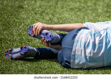 Close-up Grabbing The Foot With Sports Shoes Of A Child Soccer Player Fallen, After Suffering An Injury On The Soccer Field, Blue Uniform Clothing, Orange Soccer Cleats, Grass Blurred In Background