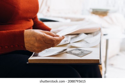 Close-up Of Grandmother Looking Old Black And White Film Photos While Sitting At Home Alone. Side View Of Female Wrinkle Hand Holding Retro Photos. Selective Focus In Foreground, Blurred Background.