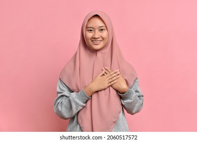 Close-up grateful, Muslim Asian woman feeling touched and flattered, smiling and gazing with affection, touch heart, express gratitude and delight of receiving nice gift, isolated on pink background