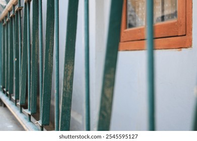 A close-up of a green metal fence with a wooden window and a gray wall in the background. The fence is made of vertical bars and is slightly rusted. Close-Up of a Green Metal Fence. - Powered by Shutterstock