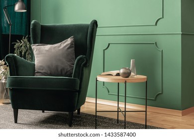 Close-up of a green velvet armchair with a gray cushion standing next to a wooden coffee table in a cozy living room interior with molding on the wall. Real photo