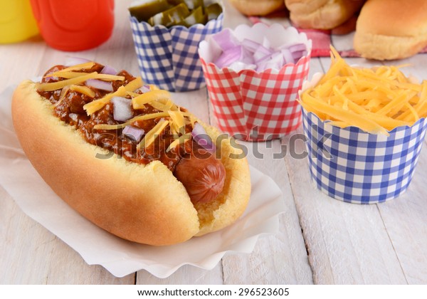 Closeup of a grilled chili dog with cheese and onions on\
a rustic wood picnic table. More buns and condiments fill the\
background. 