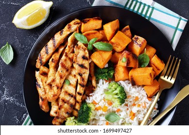 Close-up Of Grilled Paprika Chicken Strips Served With Roasted Sweet Potatoes, Rice And Broccoli On A Black Plate With Fork And Knife On A Concrete Table, View From Above, Flat Lay, Free Space