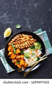 Close-up Of Grilled Paprika Chicken Strips Served With Roasted Sweet Potatoes, Rice And Broccoli On A Black Plate With Fork And Knife On A Concrete Table, View From Above, Flat Lay, Free Space