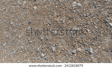 A close-up of the ground showing an array of small stones, gravel, and sandy soil. The texture is rough with a mix of neutral colors, predominantly shades of brown and grey.