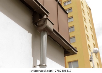 Closeup gutter and drainpipe on facade with distant apartment tower, metal brackets and joints visible, workmanlike texture, practical maintenance context