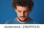 Close-up of guy with curly hair dressed in blue t-shirt stands with head down and eyes closed on blue background in the studio