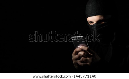 Close-up Of Hacker Using Smart Phone. Young adult girl in black clothes with hidden face looks at smartphone screen on black background with copy space.