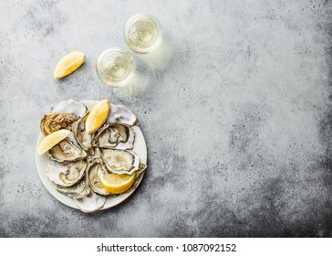 Close-up of half dozen of fresh opened oysters and shells with lemon wedges on a plate, two glasses of white wine, top view, grey rustic concrete background, space for text