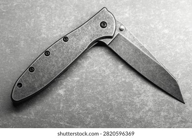 Close-up of a half opened folding pocket knife with a stonewashed blade and metal handle resting on a textured surface, conveying utility, craftsmanship and industrial minimalism