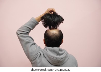 Un gros plan d'un mâle à moitié chauve enlevant sa perruque tout en portant un capuchon sur fond rose