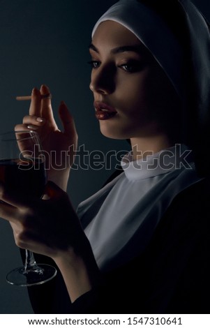 Close-up half-turn portrait of a nun, posing on a black background. She's wearing dark nun's clothing. The nun is holding a cigarette in a right hand and glass of wine in a left hand. 