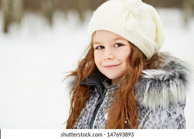 Closeup half-turned portrait of little girl in grey jacket with fur collar and knitted hat in snowy park