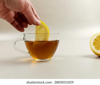 Close-up of a hand adding a fresh lemon slice into a glass cup of hot black tea on white background.