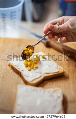 Close-up of a hand adding sweet corn kernels to white toast with a creamy spread on a wooden board. Ideal for recipes, healthy eating, or snack concepts.