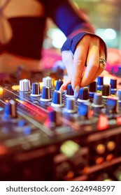 A close-up of a DJ’s hand adjusting the knobs on a vibrant, illuminated sound mixer during a live performance. The image captures the dynamic atmosphere of nightlife and professional music mixing.
