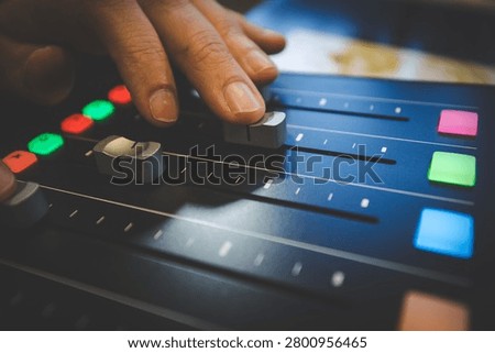 A close-up of a hand adjusting linear faders on a dark professional audio mixing console with illuminated coloured buttons, suitable for music production, broadcasting and sound engineering concepts.