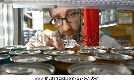 Close-up of the hand of a bearded man with glasses taking one can of soda or energy drink in a store fridge