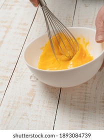 A closeup of a hand beating an egg on a bowl with an egg beater on a white plank table