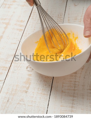 A closeup of a hand beating an egg on a bowl with an egg beater on a white plank table