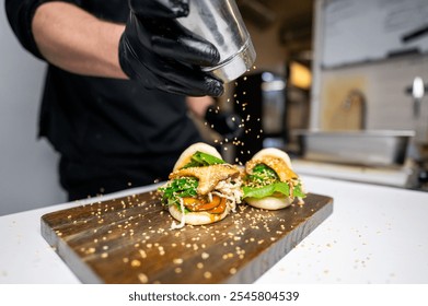 A close-up of a hand in a black glove sprinkling sesame seeds over two gourmet burgers on a wooden cutting board, with a blurred kitchen background. Perfect for food presentation and culinary themes. - Powered by Shutterstock