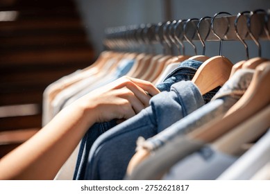 Close-up of a hand browsing through clothes on wooden hangers. The scene is well-lit, showcasing a variety of casual clothing in a retail setting.