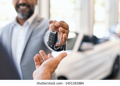 Closeup Hand Of Cardealer Giving New Car Key To Customer. Salesman Hand Giving Keys To A Client At Showroom. Man's Hand Receiving Car Keys From African Agent In A Auto Dealership With Copy Space.