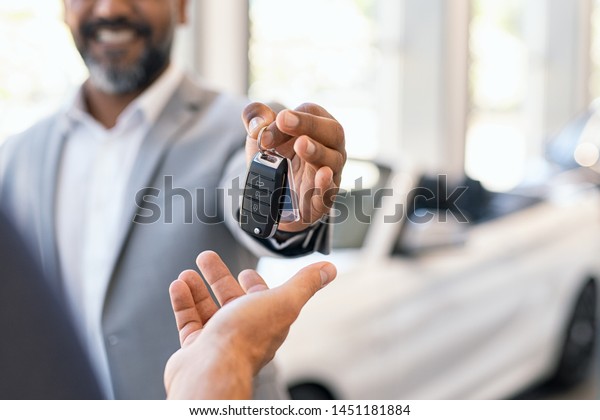 Closeup hand of cardealer giving new car key to
customer. Salesman hand giving keys to a client at showroom. Man's
hand receiving car keys from african agent in a auto dealership
with copy space.