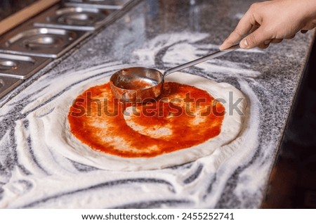 Closeup hand of chef baker making pizza at kitchen. The process of preparing pizza. Chef pours tomato sauce over the rolled dough. Chef hand is spreading pasteurized tomato paste onto a pizza base.
