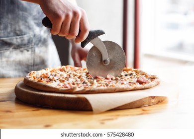 Closeup hand of chef baker in uniform blue apron cutting pizza at kitchen - Powered by Shutterstock