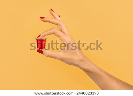 Closeup of hand of crop anonymous woman holding red coffee capsule against yellow background in studio