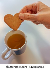 Closeup Of Hand Dipping Ikea Cookie Biscuit Into Cup Of Tea, Heart Shaped Cookie And Mug 