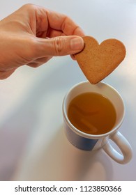 Closeup Of Hand Dipping Ikea Cookie Biscuit Into Cup Of Tea, Heart Shaped Cookie And Mug 