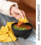 A close-up of a hand dipping a tortilla chip into a small bowl of salsa, surrounded by several chips on a wooden table.