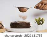 Close-up of a hand dusting cocoa powder over a chocolate cake using a fine sieve, with a minimalistic setup featuring a potted plant and kitchen utensils in the background.