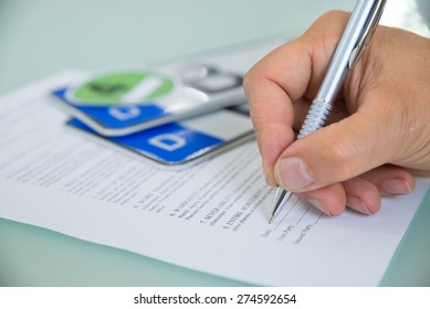 Close-up Of A Hand Filling Car Sale Contract Form With Vehicle Registration Plate On Desk. Contract Paper Contains Placeholder Text